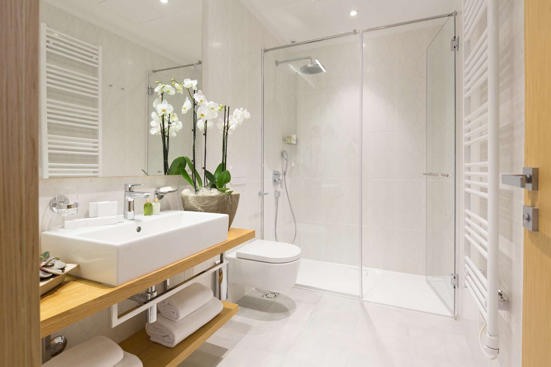 Lichte moderne badkamer met ruime inloopdouche, zwevend toilet, houten wastafelmeubel, designradiator en orchideeën – inspiratie voor een frisse en luxe badkamer inrichting.