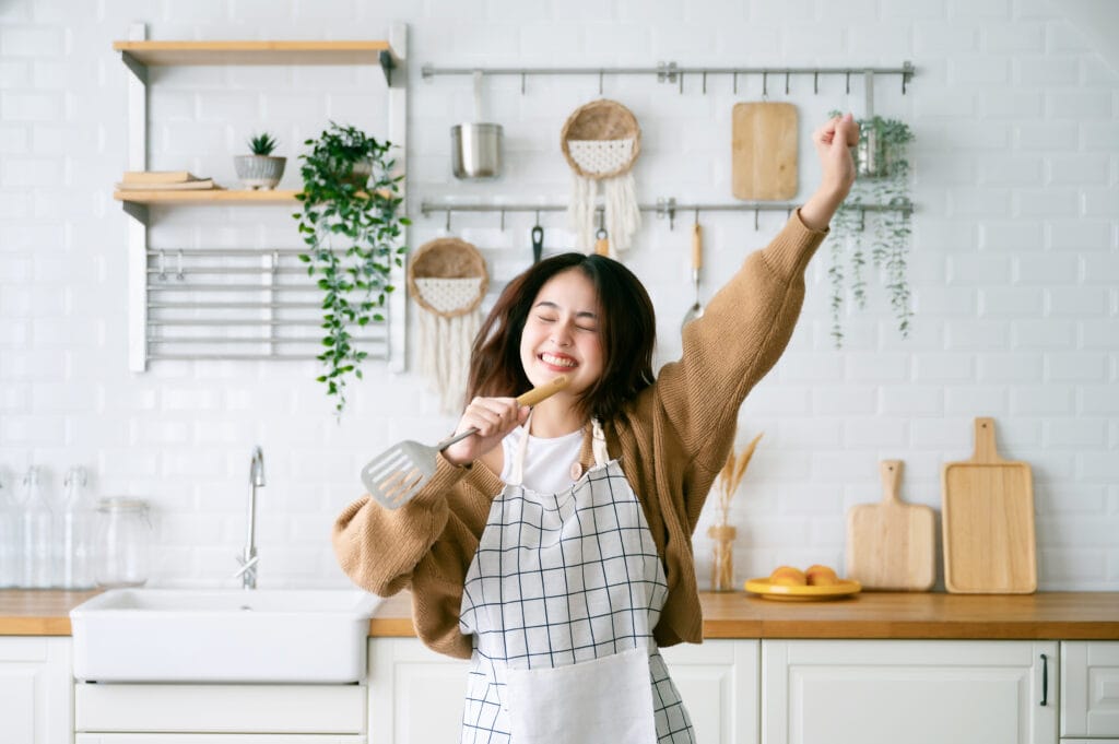 Vrolijke jonge vrouw met schort zingt en danst in een lichte, moderne keuken met witte metrotegels als achterwand. De keuken heeft houten accenten, hangende planten, en stijlvolle accessoires aan de muur. Deze gezellige sfeer maakt koken leuker en inspireert tot creativiteit in huis.