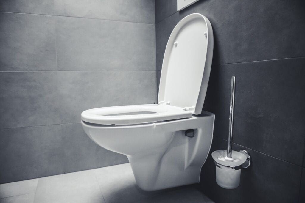 Modern wandcloset met grote grijze vloertegels en wandtegels in een minimalistisch toilet, met een toiletborstel aan de muur.