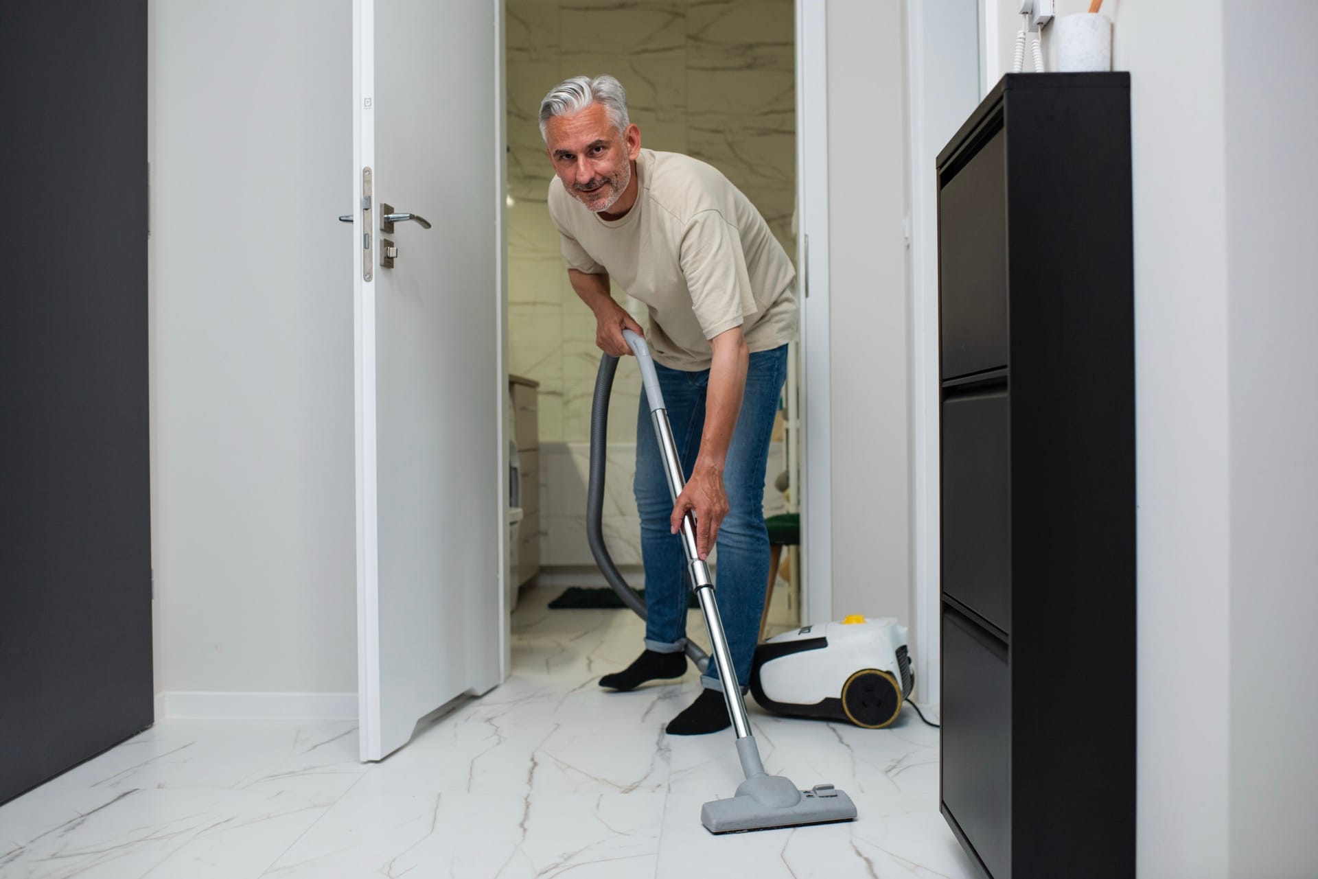 Hand die witte marmerlook tegels schoonmaakt met een gele doek – onderhoud badkamertegels, tips voor tegelreiniging, glanzende wandtegels schoonmaken voor hygiënische badkamer. Man stofzuigt de vloer in huis.