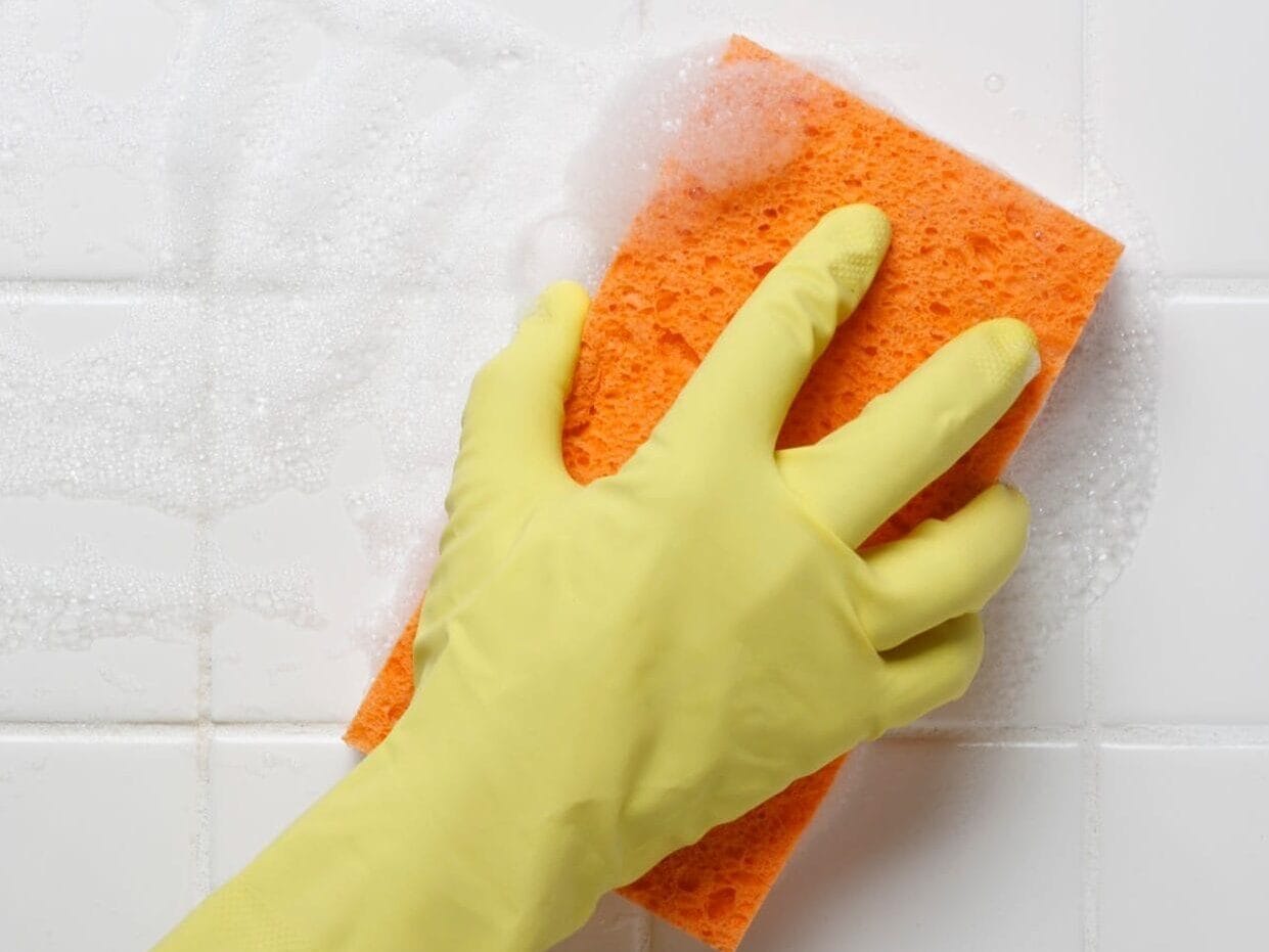 Vrouw maakt tegelvloer schoon met dweil – effectief tegelvloer reinigen, glanzende en hygiënische vloertegels in huis, beste schoonmaakmethode voor tegelvloeren. Close-up van een hand met een gele schoonmaakhandschoen die een oranje spons gebruikt om glanzende, witte badkamertegels te reinigen.
